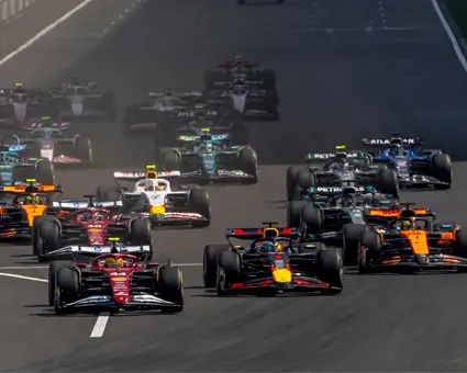 A group of Formula 1 cars racing side by side on a wide track during the start of a race, with colorful liveries and a safety car visible in the distance.