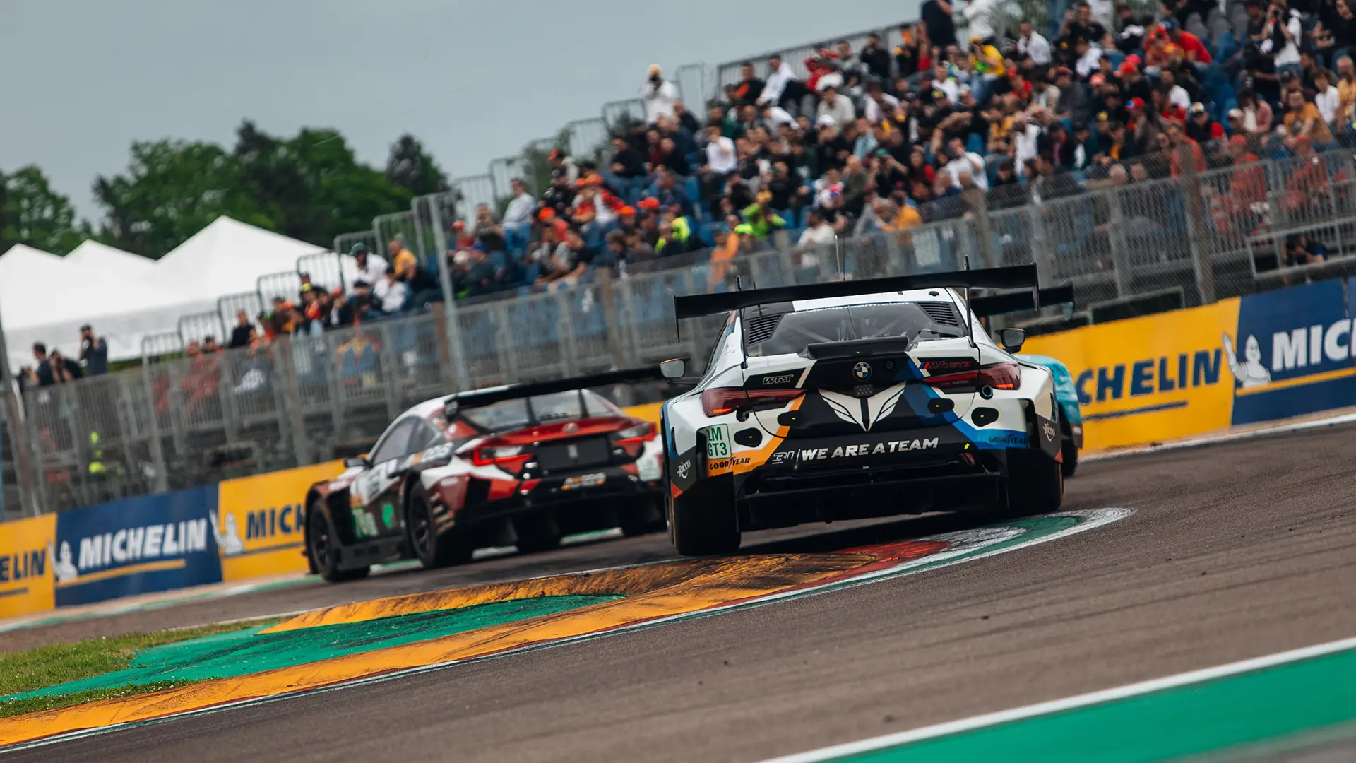Two GT race cars speeding on a track with colorful curbs, viewed from behind, as a crowd watches from grandstands lined with Michelin banners.