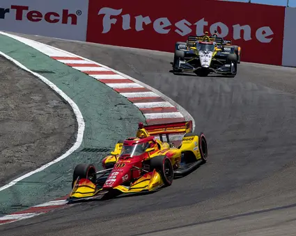 A red and yellow IndyCar leading through a sharp uphill turn on a racetrack, followed by a black and yellow car, with sponsor banners for WeatherTech, Firestone, and Java House visible on the barriers.