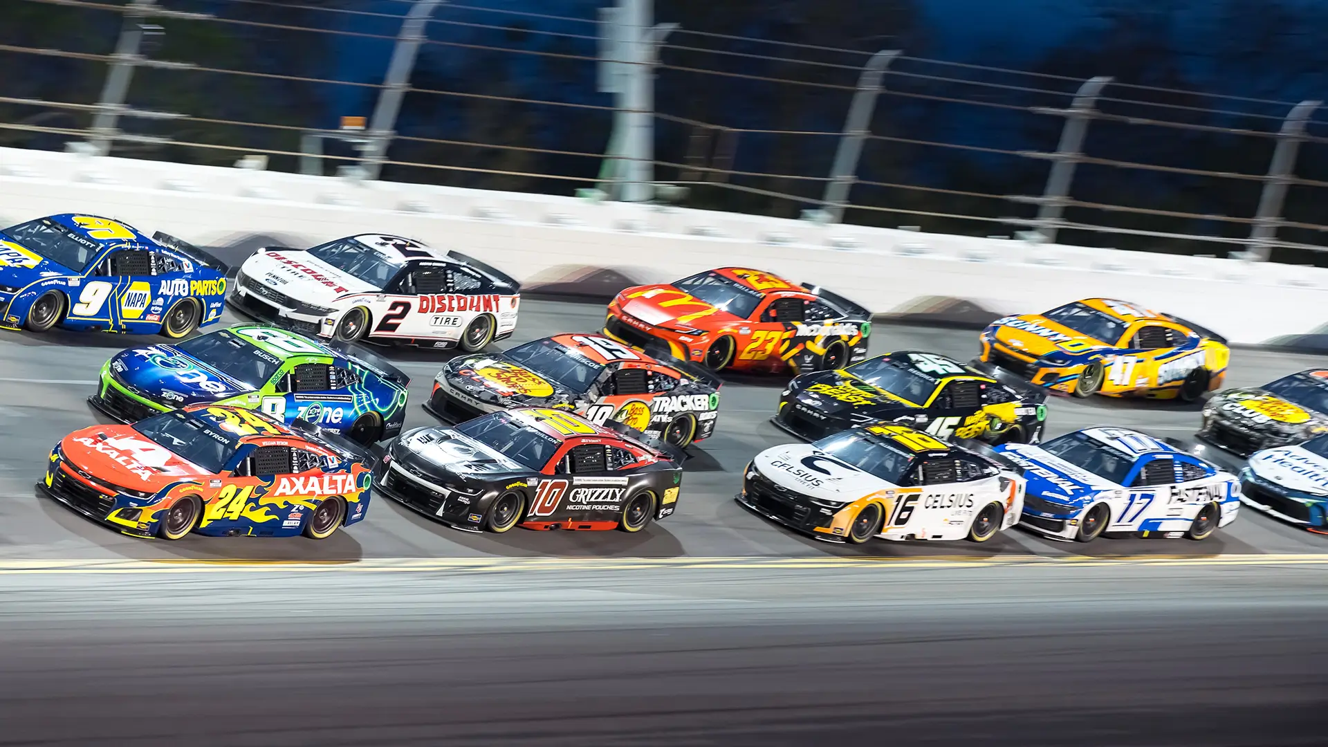 A pack of brightly colored stock cars racing side by side on a banked oval track under stadium lights, with safety barriers lining the track.
