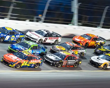 A pack of brightly colored stock cars racing side by side on a banked oval track under stadium lights, with safety barriers lining the track.