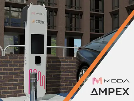 A sleek black electric car parked next to a white ultra-rapid EV charging station branded with “MODA” and “Motion Applied.” The charger has a digital display and pink cable graphics at the bottom. The setting is an urban area with a brick wall and modern office building in the background.