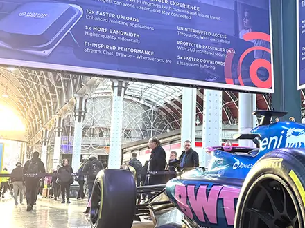 A blue and pink Alpine Formula 1 race car displayed indoors at a train station concourse, with large promotional banners overhead advertising high-speed Wi-Fi features. Sunlight streams through the glass roof, and people walk around the exhibit in the background.