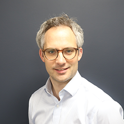 Harry Lowe, Director, Finance, a light-colored button-down shirt, posed against a plain dark gray background in a formal portrait style.