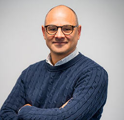 Richard Saxby, Director, Motorsport, wearing a navy blue cable-knit sweater over a light patterned collared shirt, posed with arms crossed against a plain light gray background.