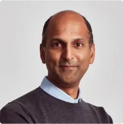 Samir Maha, Chief Executive Officer, middle-aged with very short dark hair smiling, wearing a grey jumper over a light blue shirt.