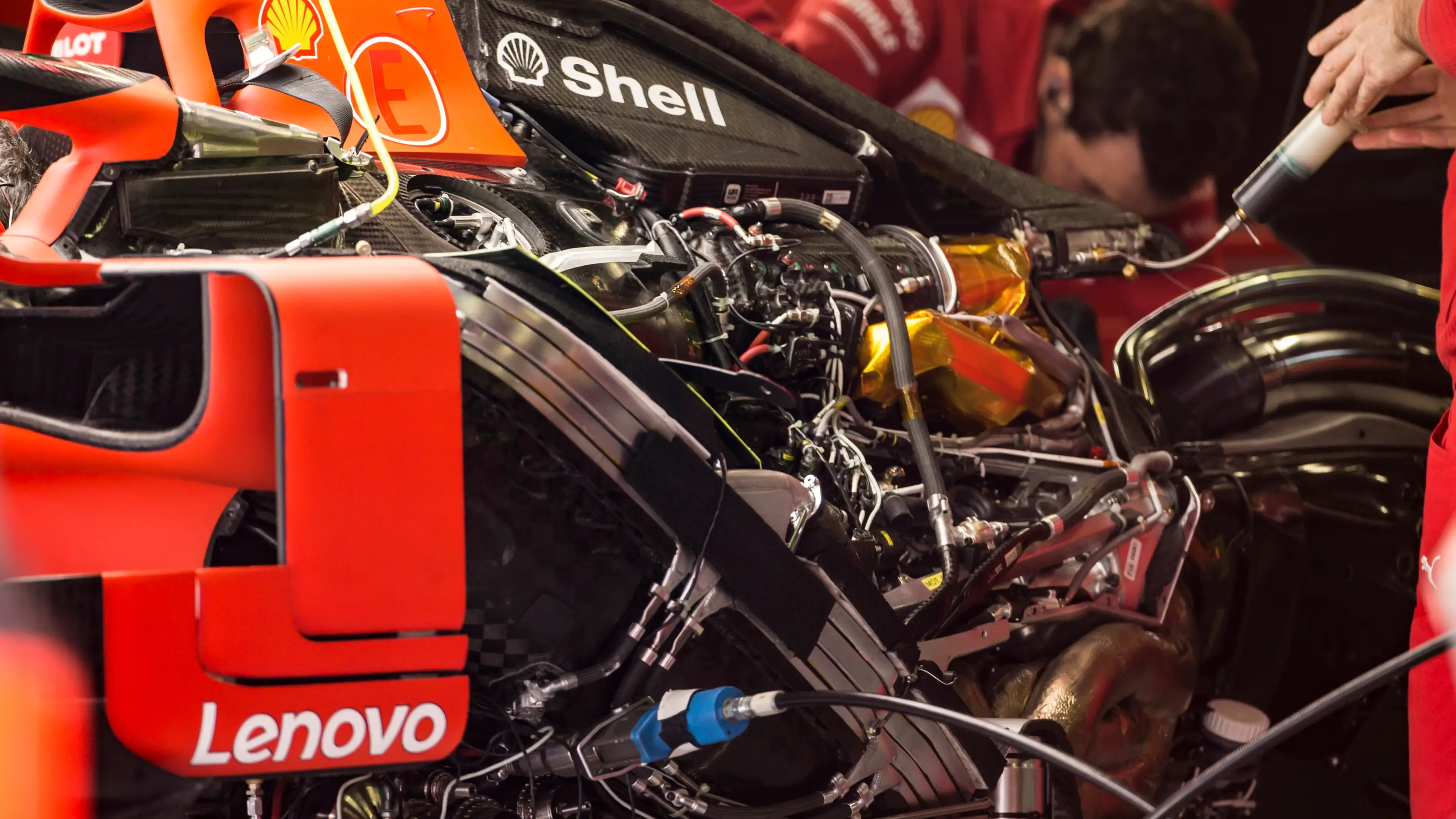 Close‑up of a Formula One race car engine bay, showing exposed mechanical components, wiring, and fluids as a team member works on the car.