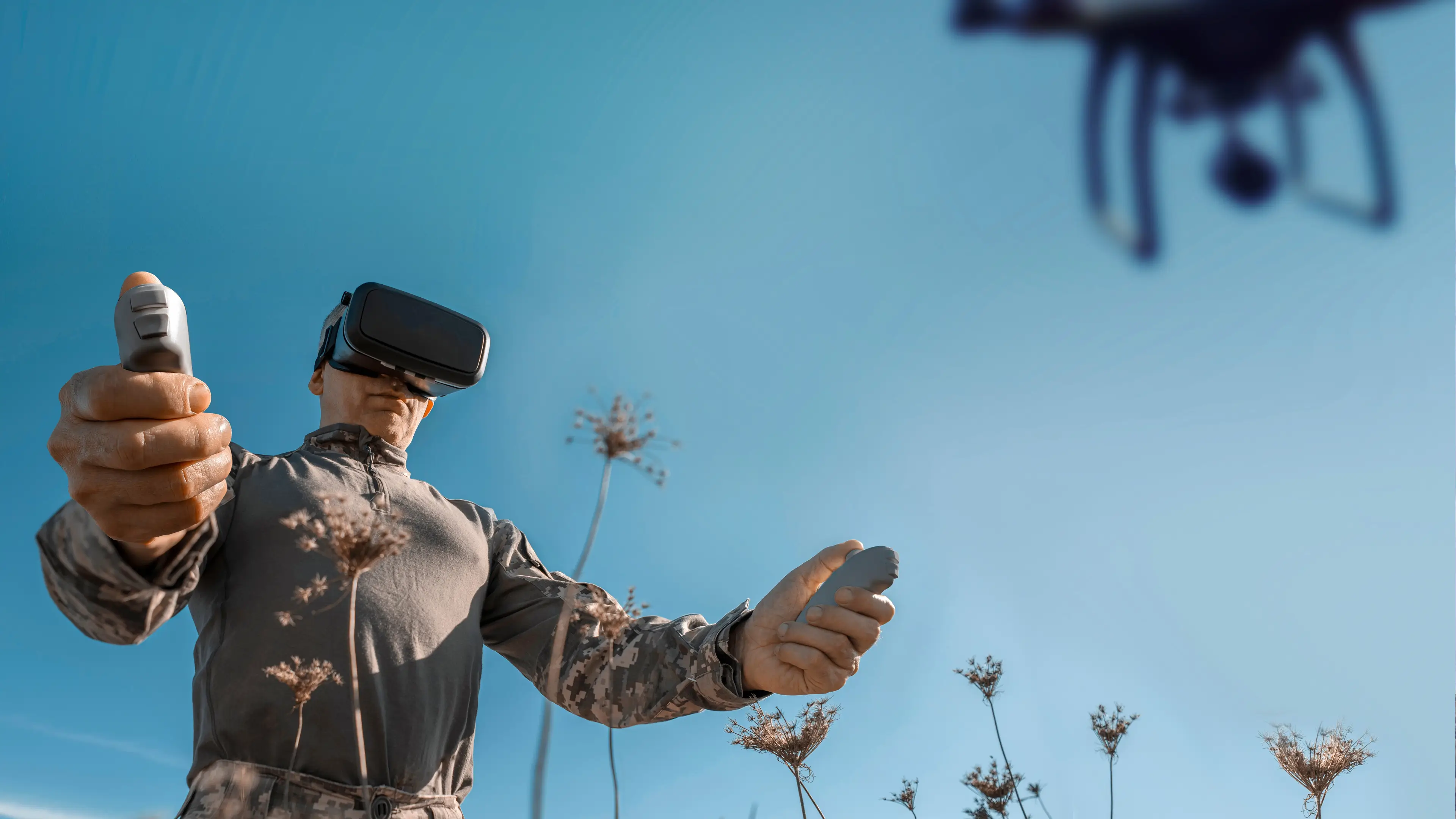 Person wearing a virtual reality headset and holding handheld controllers while operating a drone outdoors against a clear blue sky.