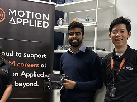 Group of people standing in a Motion Applied office or lab, with Motion Applied branding visible on banners and a screen, and one person holding an electronic control unit in front of display shelves