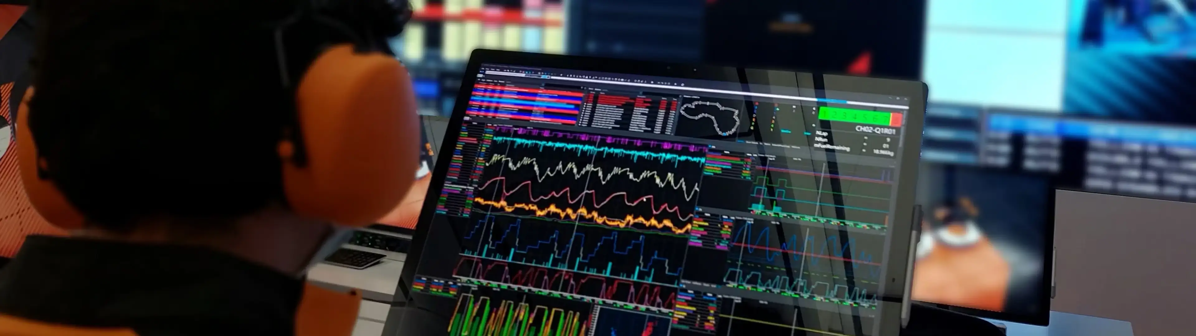 Person wearing over‑ear hearing protection seated at a workstation, viewing multiple computer screens displaying colorful real‑time data charts and graphs in a control or monitoring environment.