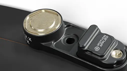 Compact Motion Applied electronic sensor module mounted on a black bracket, featuring a circular metal cap, a small rectangular housing with Motion Applied branding, and a mounting hole, shown against a dark background.