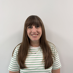 Lynette Prag, Director, HR, with long, straight brown hair wearing a white T-shirt with green horizontal stripes, standing against a plain light-colored wall.