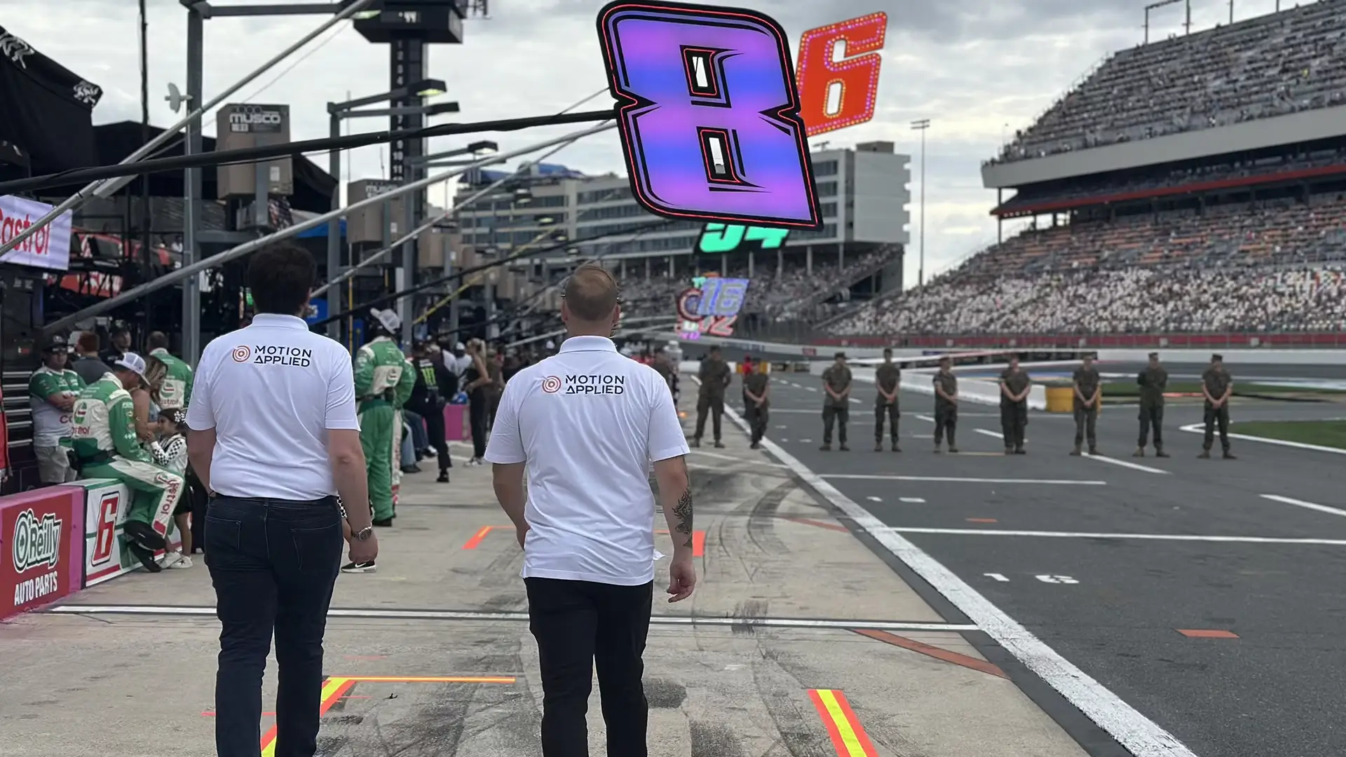 Pit lane scene at a motorsport race, with two team members wearing Motion Applied shirts walking toward the track as race cars, pit equipment, and grandstands filled with spectators appear in the background.