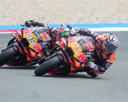 Two motorcycle racers leaning sharply into a corner on a racetrack, riding closely side by side on orange racing bikes during a high‑speed turn.