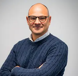 Richard Saxby, Director, Motorsport, wearing a navy blue cable-knit sweater over a light patterned collared shirt, posed with arms crossed against a plain light gray background.