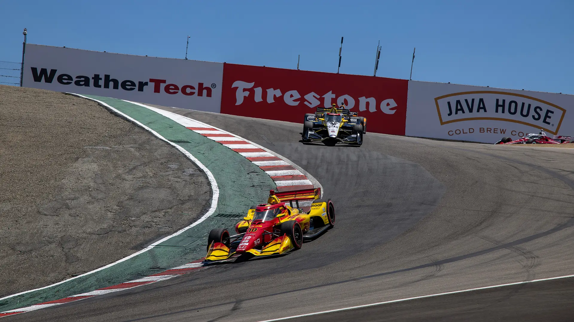 Two INDYCARs race down the corkscrew at Laguna Seca Raceway, with a red and yellow car leading and another car following.