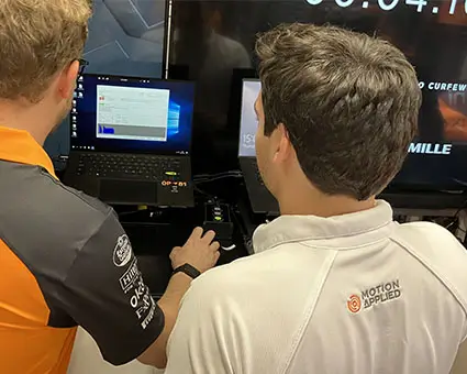 Two people viewed from behind using a laptop and control hardware at a Motion Applied workstation, with software displayed on the screen during a hands‑on training or demonstration.