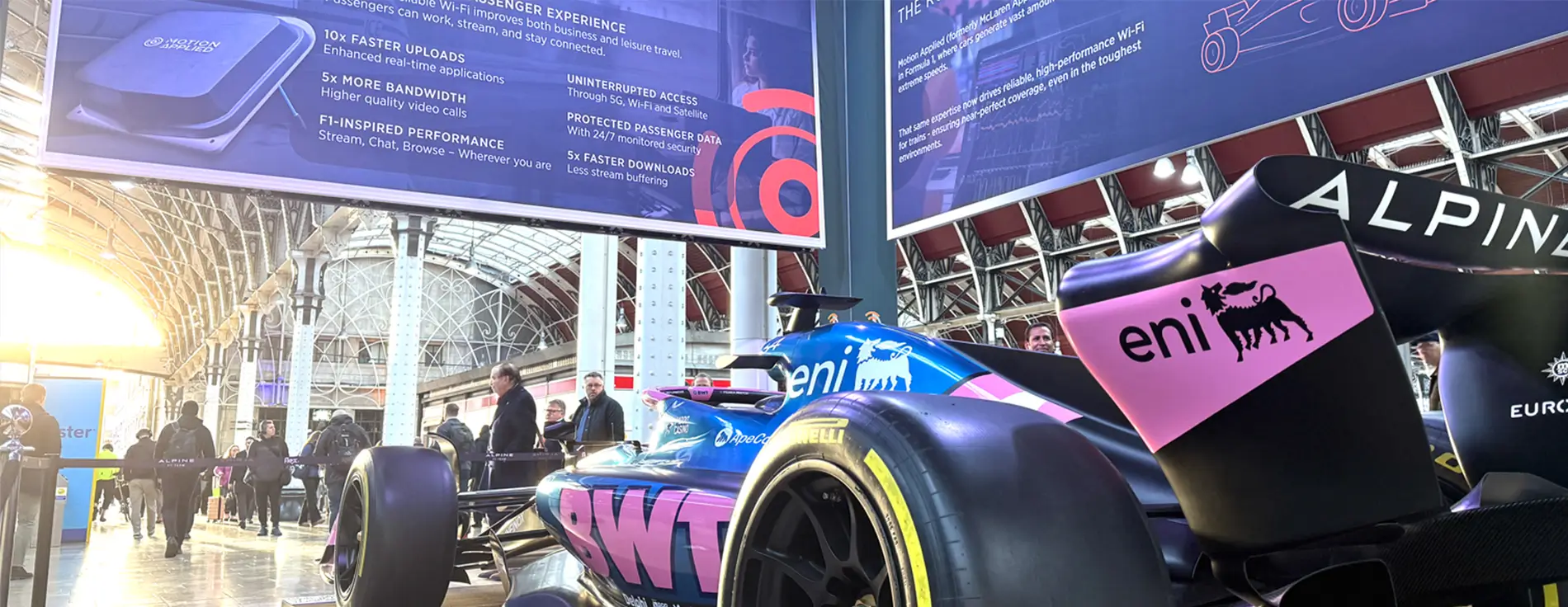 Formula‑style race car displayed indoors at an exhibition, viewed from the rear side, with people walking nearby and large overhead banners showing connectivity and performance information.
