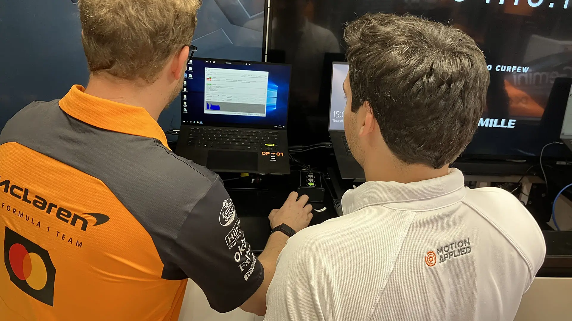 Two people viewed from behind using a laptop and control hardware at a Motion Applied workstation, with software displayed on the screen during a hands‑on training or demonstration.