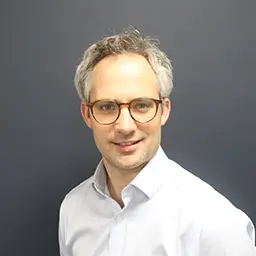 Harry Lowe, Director, Finance, a light-colored button-down shirt, posed against a plain dark gray background in a formal portrait style.