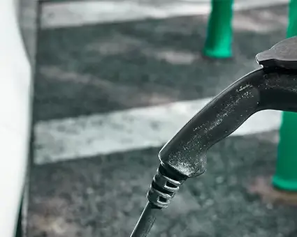 Close‑up of an electric vehicle charging connector plugged into a white car, with part of the headlight visible in an outdoor parking area.
