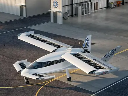 White electric aircraft with multiple rotors mounted along its wings, parked on an airport apron and viewed from above at an angle.