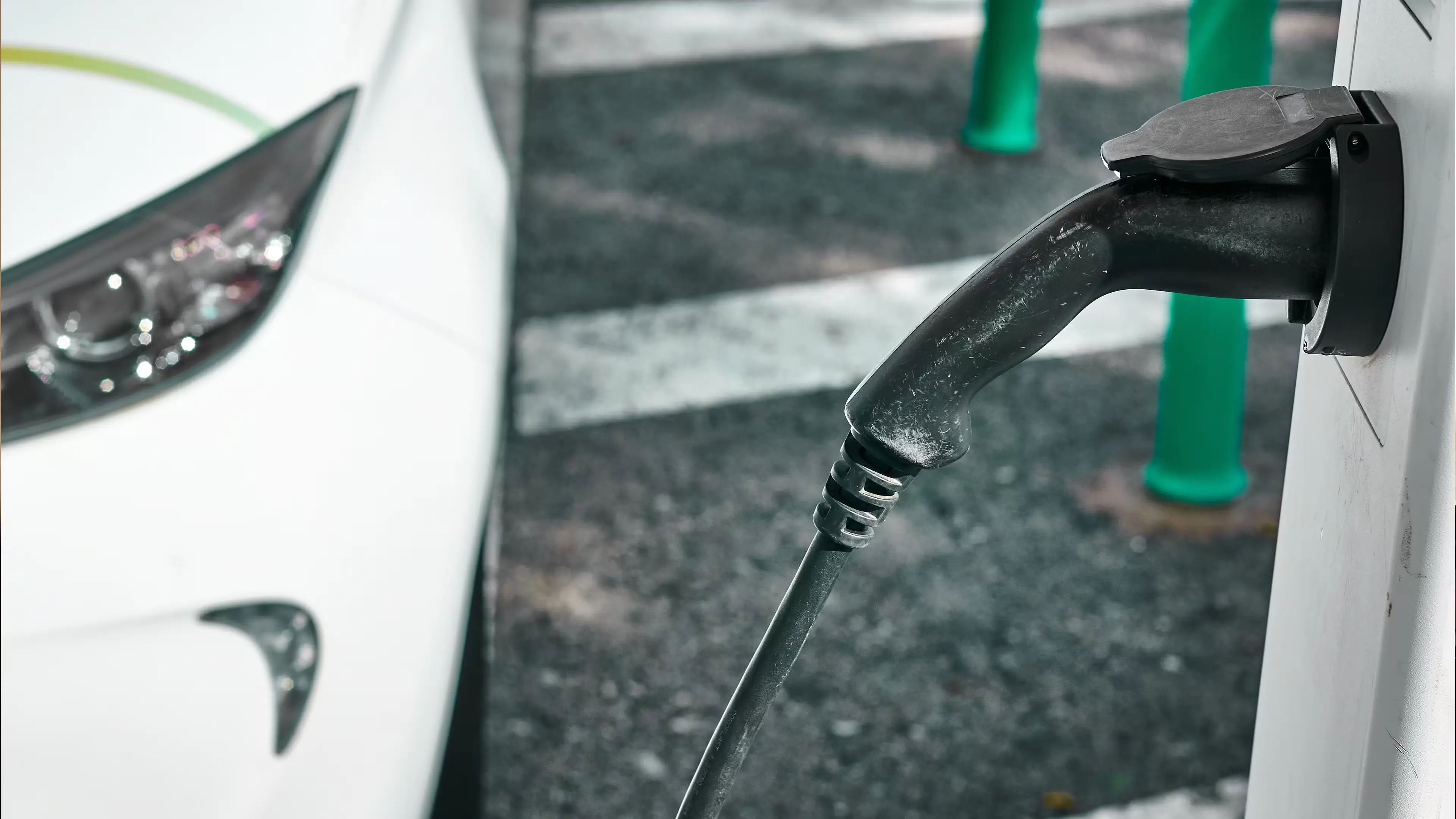 Close‑up of an electric vehicle charging connector plugged into a white car, with part of the headlight visible in an outdoor parking area.