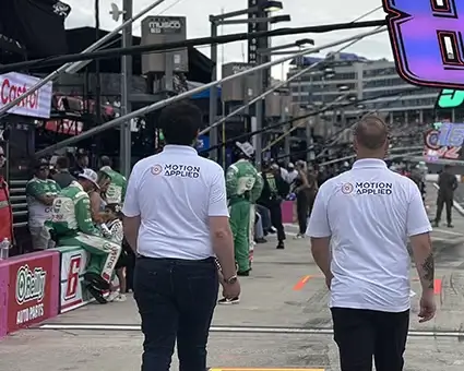 Pit lane scene at a motorsport race, with two team members wearing Motion Applied shirts walking toward the track as race cars, pit equipment, and grandstands filled with spectators appear in the background.