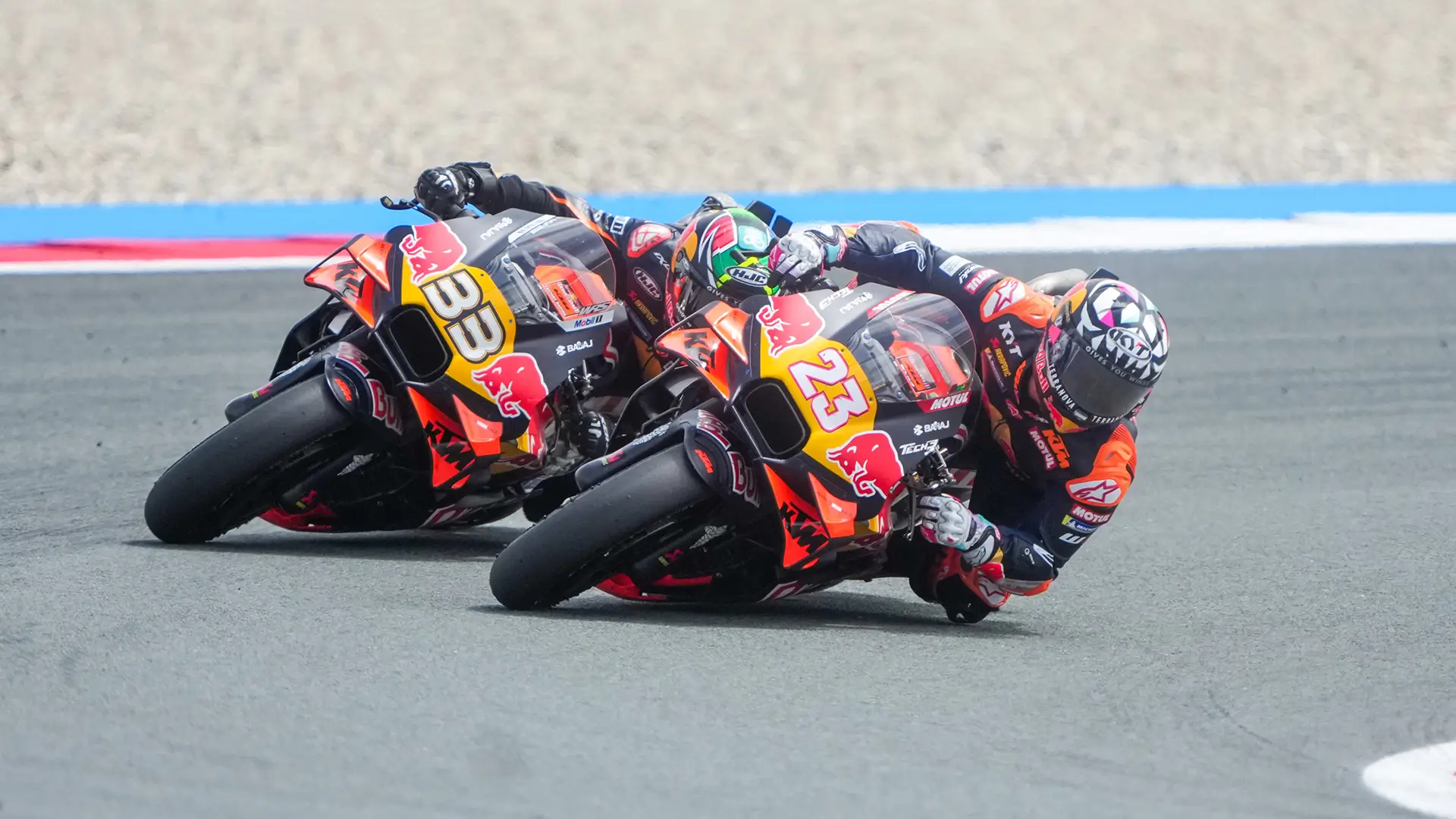 Two motorcycle racers leaning sharply into a corner on a racetrack, riding closely side by side on orange racing bikes during a high‑speed turn.