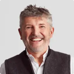 Nick Fry, Non-Executive Chairman. A middle-aged man with short styled light brown hair, smiling, wearing a white shirt undone at the top and a grey Gilet style jacket.