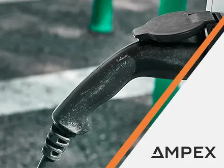 Close‑up of an electric vehicle charging connector plugged into a charging unit, shown against an outdoor parking surface with AMPEX branding visible.