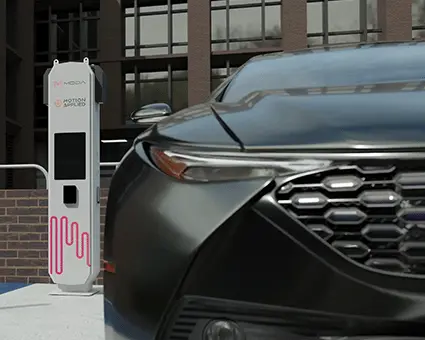 Urban street scene showing the front of a dark car parked beside a Motion Applied vehicle charging station, with the charger unit visible against a modern brick building.
