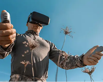 Person wearing a virtual reality headset and holding handheld controllers while operating a drone outdoors against a clear blue sky.
