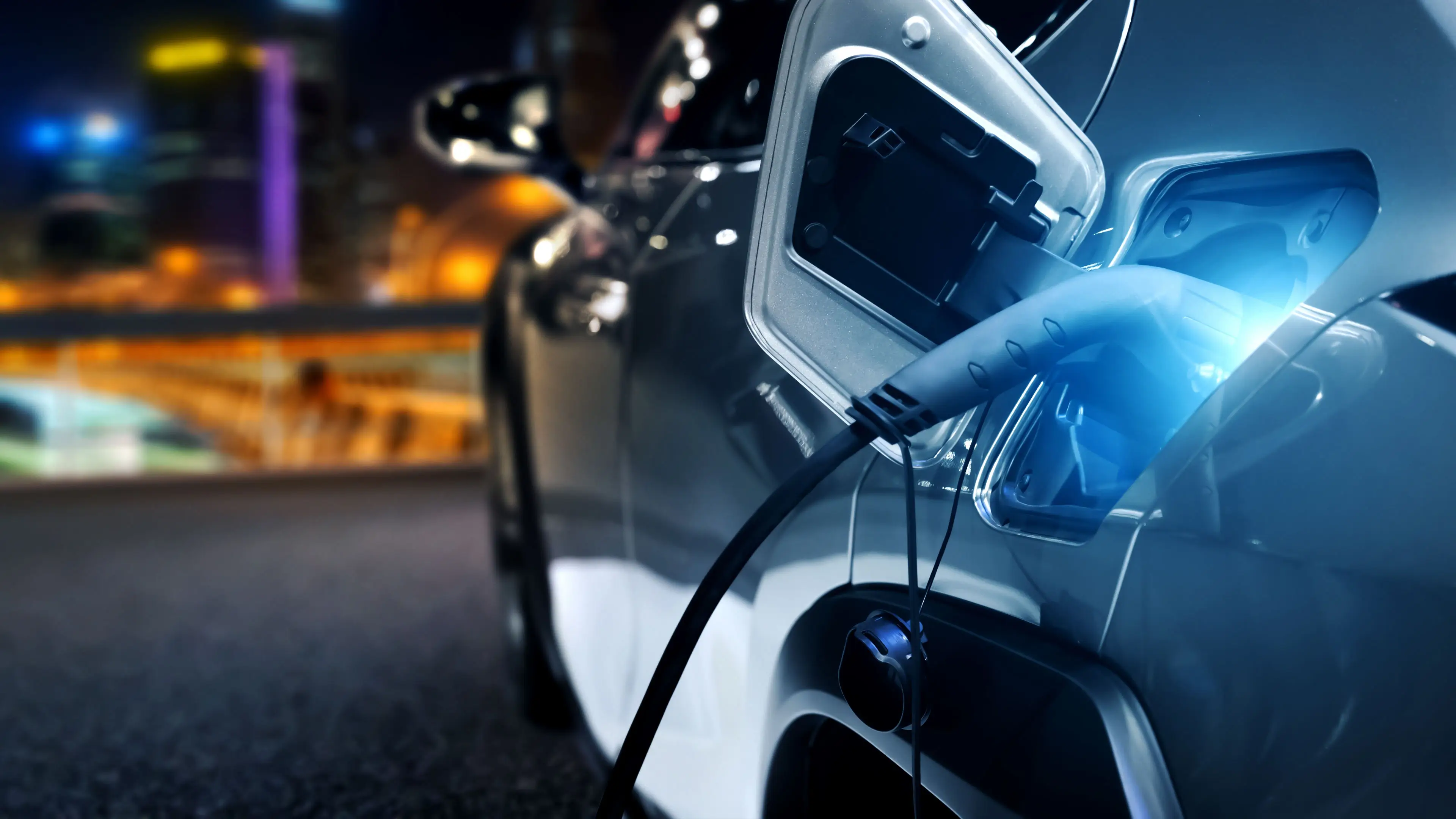 Person standing beside an electric car while using a smartphone, with an electric vehicle charging cable connected to the car in an outdoor parking area.