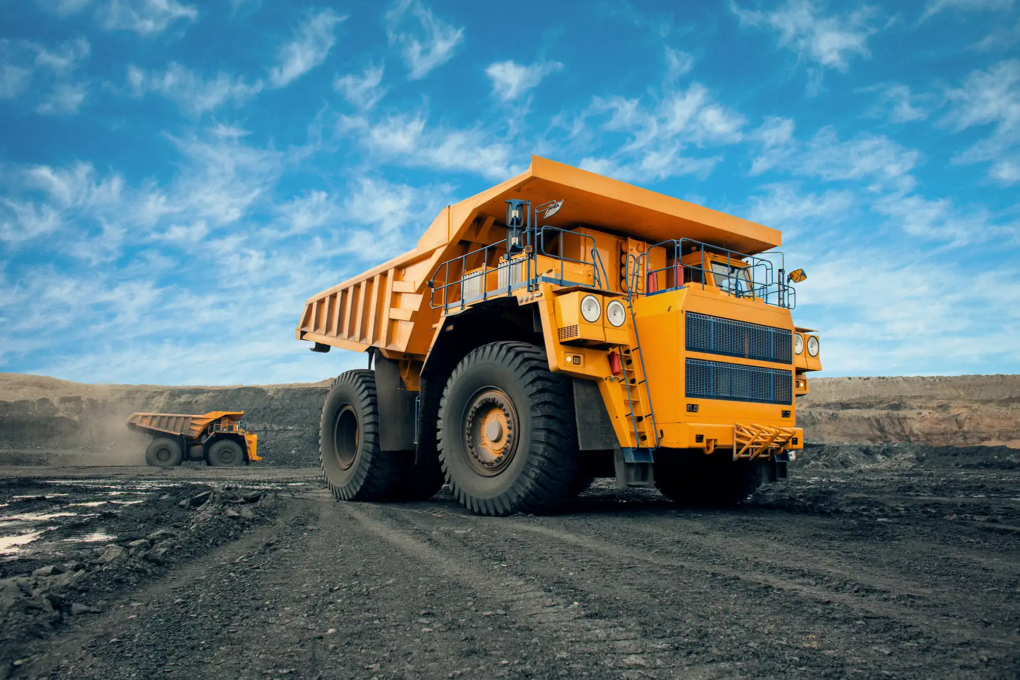 Large yellow mining haul truck driving on a dirt road in an open‑pit mine, with another haul truck visible in the background under a blue sky.