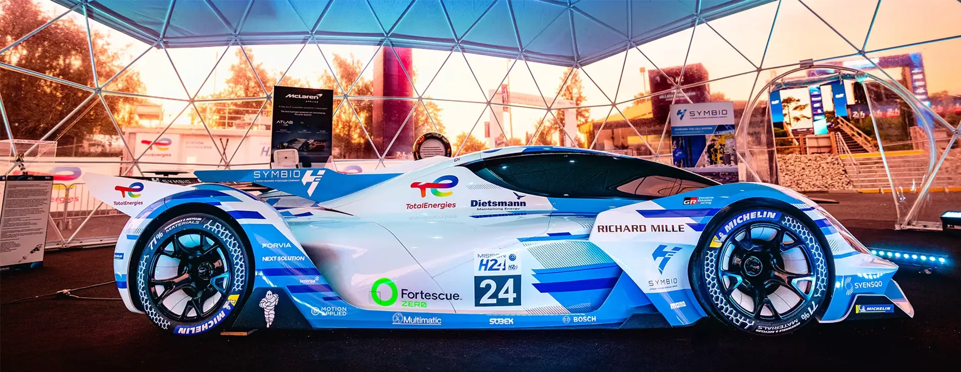 Side view of a futuristic endurance race car with blue and white livery and number 24, displayed indoors under a geodesic exhibition structure with sponsor logos visible.