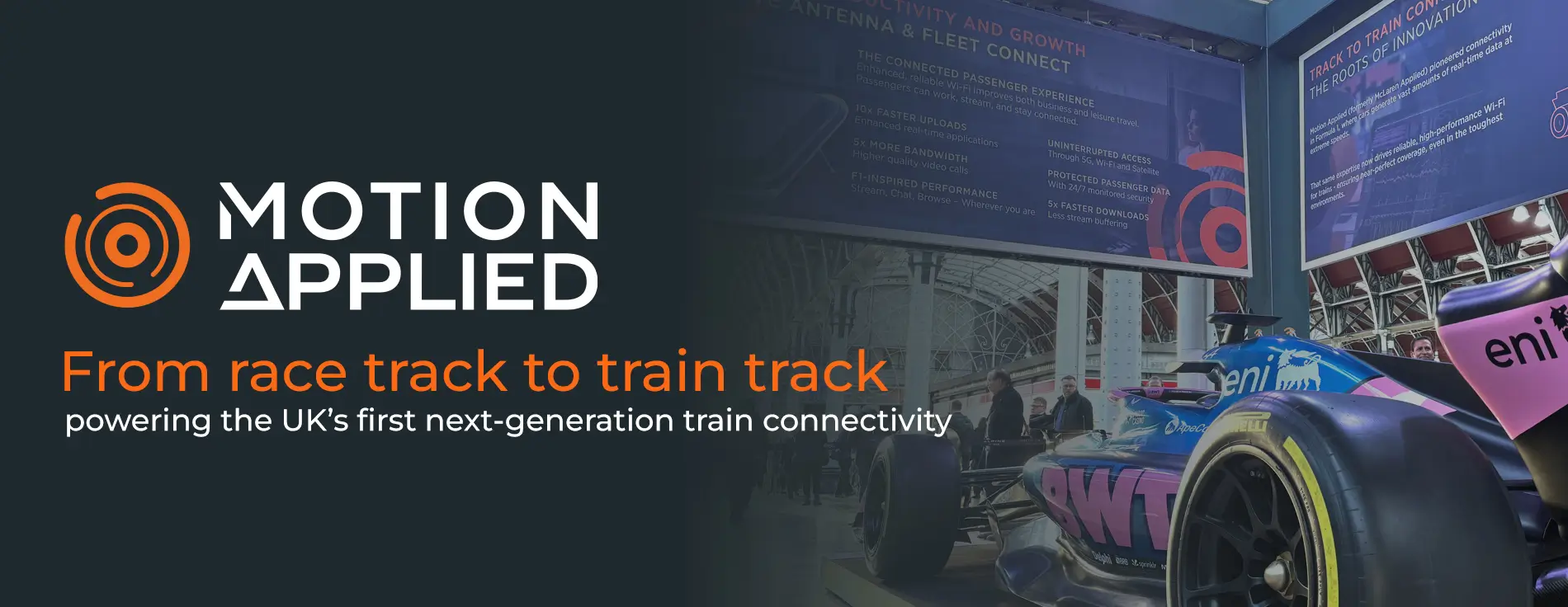 Motion Applied branding alongside a Formula‑style race car displayed indoors, with overlaid text reading ‘From race track to train track, powering the UK’s first next‑generation train connectivity,’ and exhibition displays visible in the background.