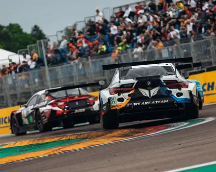 Two GT‑style race cars viewed from the rear as they corner on a racetrack, with spectators watching from grandstands in the background.