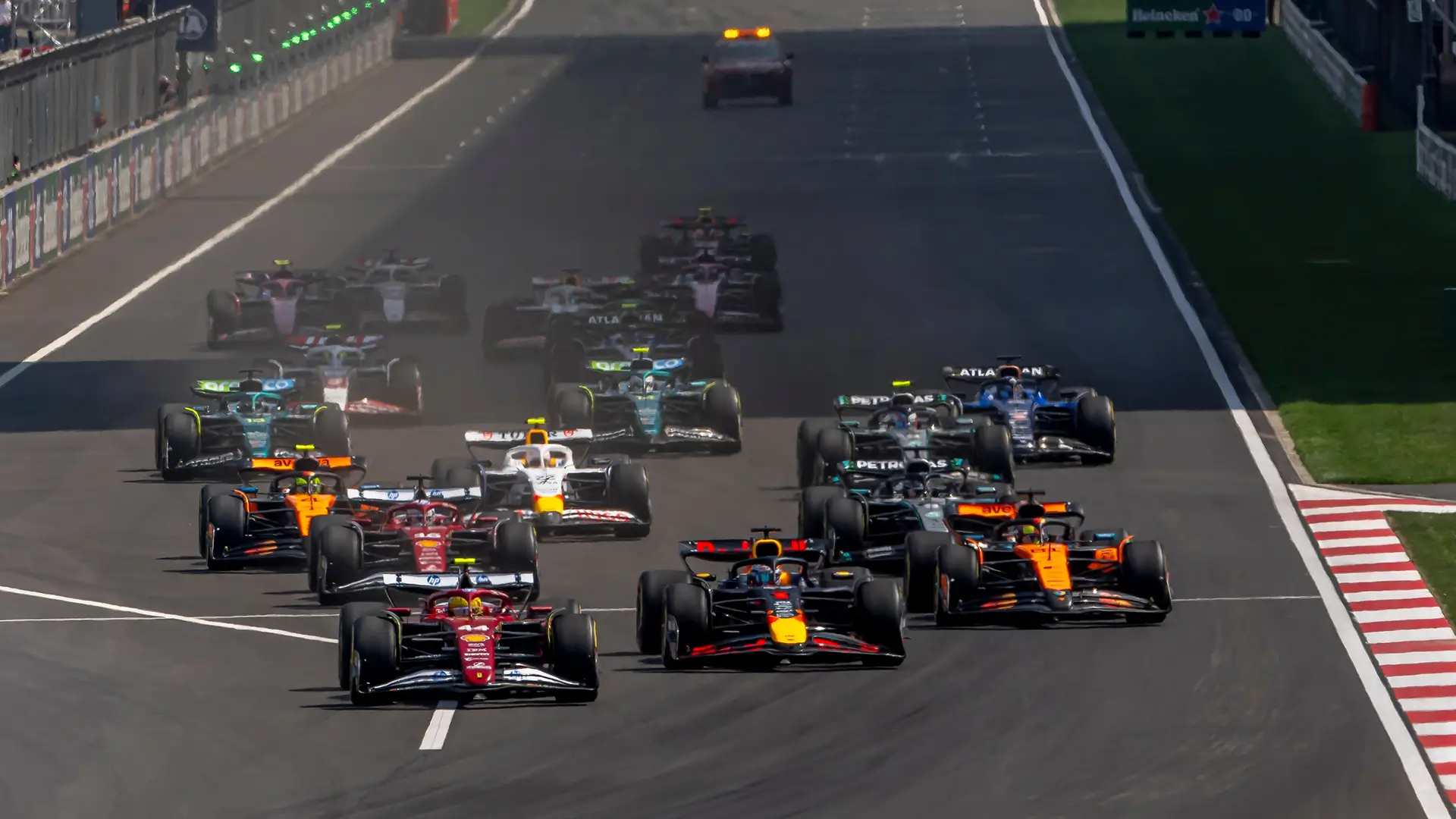 Group of Formula‑style race cars accelerating closely together on a racetrack, viewed from the front as they approach a straight section.